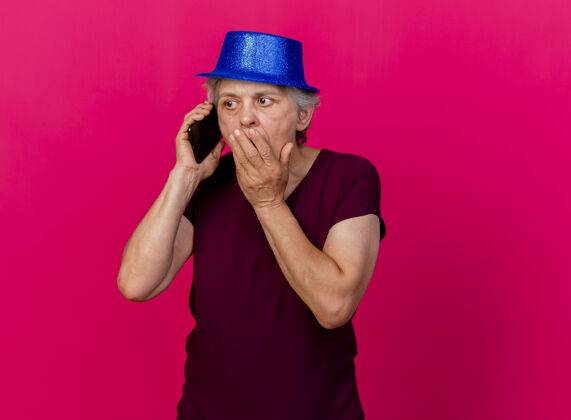 聚会震惊的老妇人戴着聚会的帽子把手放在嘴上讲电话就粉红色电话女人手