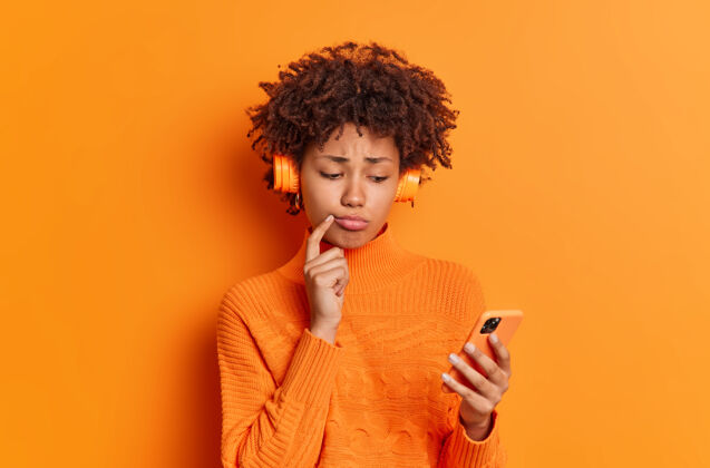 担心心烦意乱的千禧一代女孩沮丧的表情读短信听音乐播放列表穿着休闲毛衣隔离在生动的橙色墙壁痛苦美丽科技