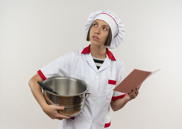 感觉体贴的年轻女厨师穿着厨师制服 拿着锅和便笺簿 孤零零地看着白色的墙上白拿着垫