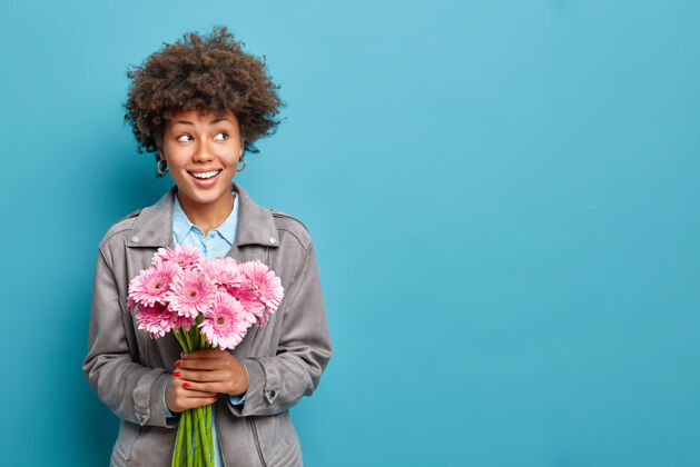 美丽可爱开朗的女人拿着一束粉红色非洲菊 穿着灰色夹克模特在蓝色的墙上庆祝春节春天欢呼情绪