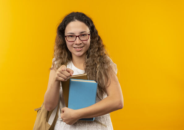 女生微笑着的年轻漂亮的女学生戴着眼镜 背着书包 指着隔离在黄色墙上的前面市民姿势背