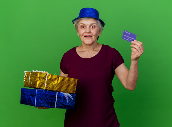 盒子戴着派对帽的兴奋的老妇人拿着礼品盒和信用卡在绿色的草地上女人帽子兴奋