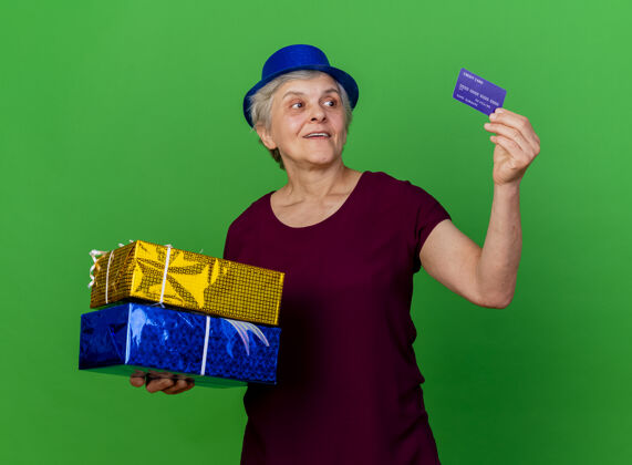 老人一位戴着派对礼帽的惊讶的老太太拿着礼盒 看着绿色的信用卡盒子信用聚会