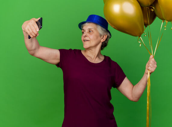 拜托高兴的老太太戴着聚会帽手持氦气球看着绿色的手机老人氦气派对