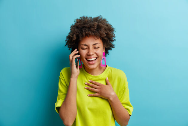 牙关无忧无虑的放松积极的非裔美国妇女一边笑一边通过智能手机说话手放在胸前闭上眼睛表达积极的情绪听到有趣的笑话穿着随便站在室内美丽说话手机