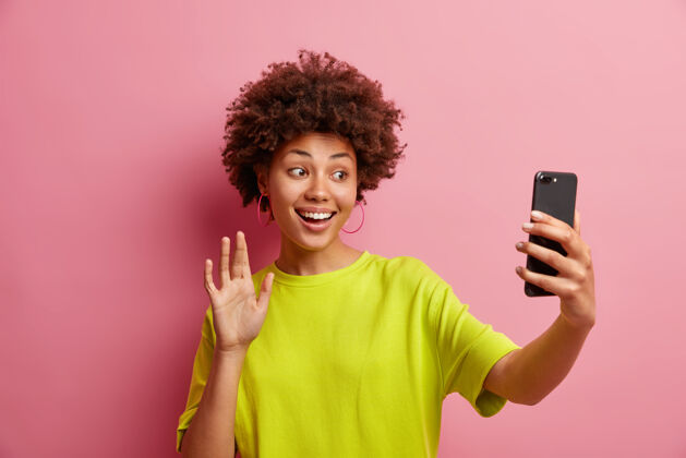 女人快乐的非洲裔美国年轻女子 在智能手机摄像头里卷起一头毛茸茸的卷发 一边打招呼 一边和远方最好的朋友进行视频会议 一边穿着休闲t恤在室内摆姿势墙视频自拍