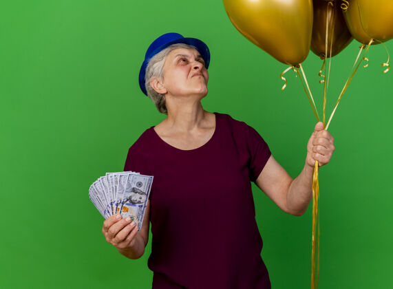 氦戴着派对帽的老妇人拿着钱 看着绿色的氦气球女人派对气球