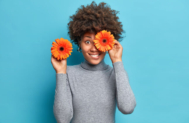 有趣积极的女人拿着橙色非洲菊 用最喜欢的花遮住眼睛 穿着休闲的灰色高领套头衫 隔着蓝色的墙女性站立乐观
