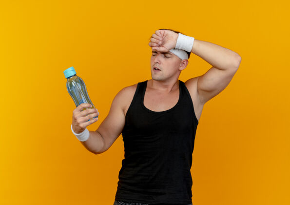 人疲惫的年轻帅气的运动型男人戴着头带和腕带 手放在额上 手拿着水瓶 看着孤立在橙色墙上的水瓶感觉姿势人