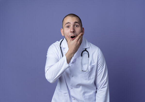 听诊器年轻的男医生穿着医用长袍 手持听诊器 下巴被隔离在紫色的墙上表情脸姿势