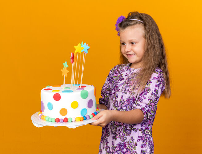 金发微笑着的金发小女孩抱着生日蛋糕 看着隔离在橙色墙上的复制空间女孩蛋糕小