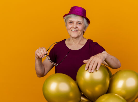 帽子微笑着戴着派对帽的老妇人站在那里 手里拿着氦气球 手里拿着隔离在橙色墙上的眼镜氦微笑光学