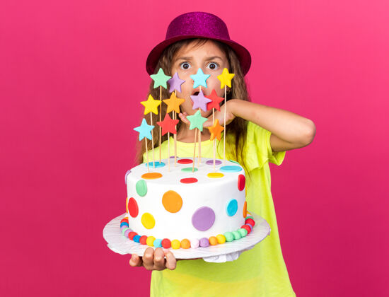 下巴惊讶的小白种女孩 戴着紫色派对帽 拿着生日蛋糕 把手放在下巴上 隔离在粉红色的墙上 留着复制空间小手女孩