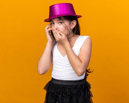 孤独震惊的年轻白人女孩戴着紫色派对帽 手放在嘴上讲电话 隔离在橙色的墙上 留着复印空间年轻帽子电话