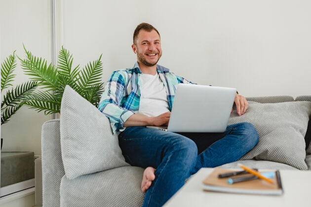 设备穿着衬衫的微笑男人在家里的沙发上放松地坐在桌子旁 在家里用笔记本电脑上网人企业家工人