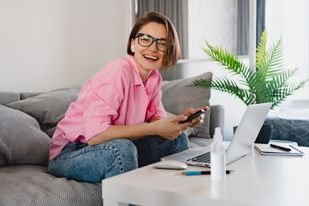 工作一个穿着粉色衬衫的微笑女人 在家里的沙发上轻松地坐在桌子旁 在家里用笔记本电脑上网房间坐姿企业家