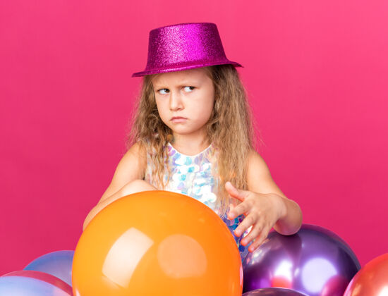 孤立恼怒的金发小女孩 戴着紫色的派对帽 站在氦气球旁 看着粉色墙壁上孤立的一面 还有复制空间小站恼火