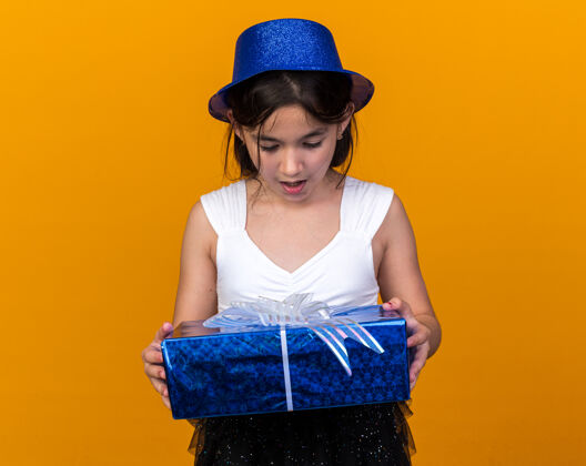 年轻惊讶的年轻白种人女孩拿着蓝色的派对帽 看着隔离在橙色墙上的礼品盒 还有复制空间帽子抱着惊讶