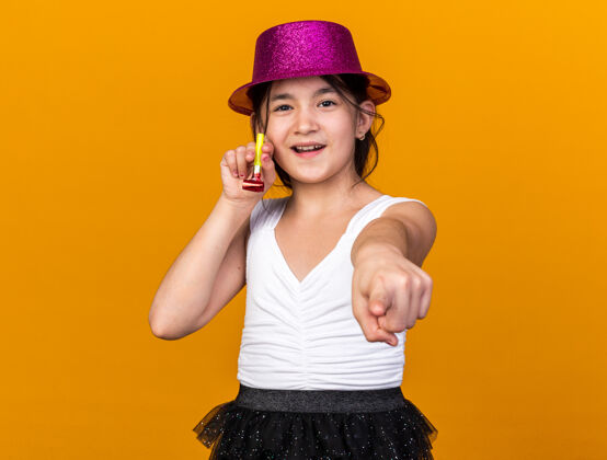 白种人高兴的年轻白人女孩 戴着紫色的派对帽 手持派对哨子 孤立地指着橙色的墙上 留着复印空间女孩孤立的拜托