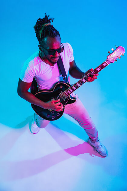 玩年轻的非洲裔美国音乐家在霓虹灯下像摇滚明星一样在蓝色的墙上弹吉他乐器球员吉他