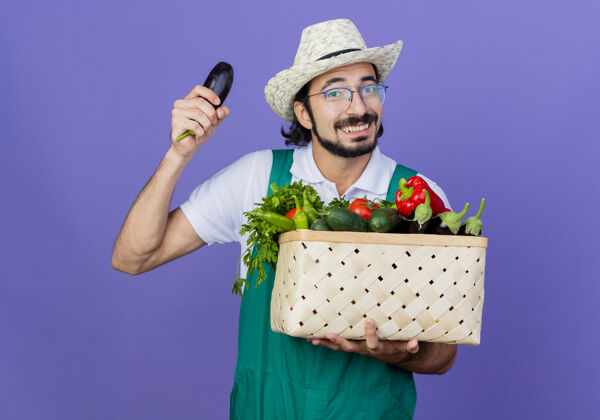 年轻人年轻的留着胡须的园丁 穿着连体衣 戴着帽子 手里拿着装满蔬菜的箱子 站在蓝色的墙上 茄子欢快地笑着表演人表情
