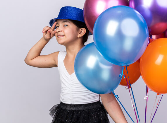 氦微笑着的年轻白种人女孩 戴着蓝色派对帽 手里拿着氦气球 打着胜利的手势 隔离在白色墙壁上 留有复制空间抱着白种人手