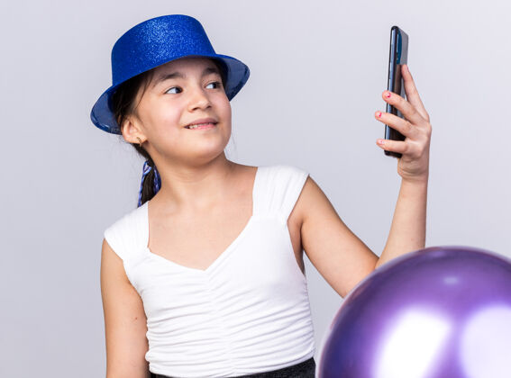 氦高兴的年轻白人女孩戴着蓝色的聚会帽站在氦气球看电话隔离在白色墙上的复制空间站着年轻孤立