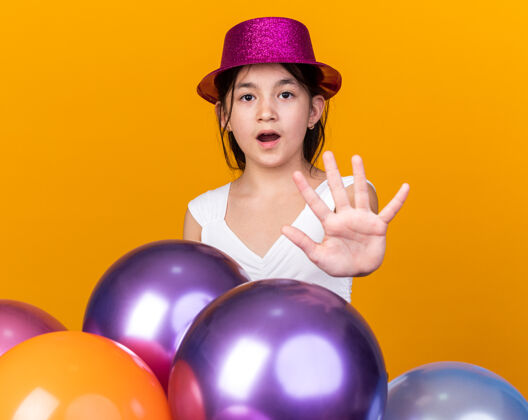 气球焦急的年轻白人女孩 戴着紫色的派对礼帽 打着停车牌 站在橙色墙上的氦气球上 留着复制空间女孩孤立手势
