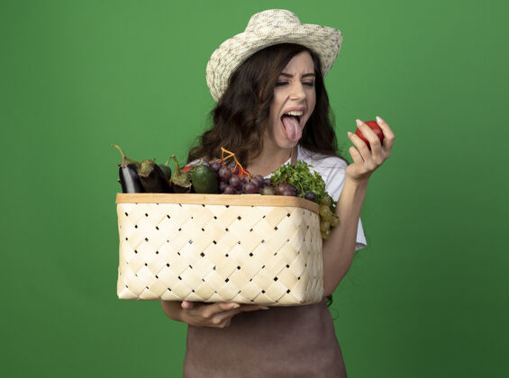 人年轻的女园丁穿着制服 戴着园艺帽 伸出舌头 把菜篮子和西红柿隔离在绿色的墙上不愉快女人蔬菜
