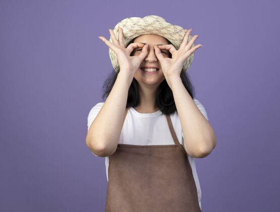 女人微笑着的年轻黑发女园丁穿着制服戴着园艺帽透过隔离在紫色墙上的手指看着前方表情脸园艺