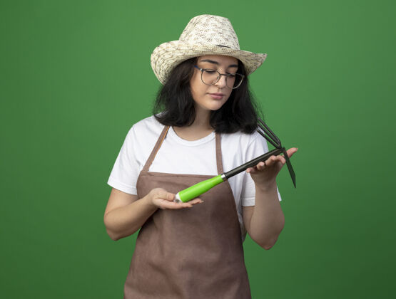 姿势自信的年轻黑发女园丁戴着眼镜和制服 戴着园艺帽 站在绿色的墙上看着锄头耙穿人园艺