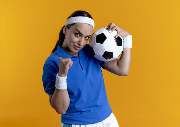 背景年轻自信的白人运动女性戴着头带和腕带拿着球指向后面球橙色背部