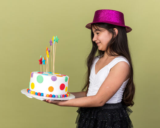 白种人微笑着的年轻白人女孩 戴着紫色的派对帽 看着生日蛋糕 站在橄榄绿的墙上 旁边有复制空间抱着孤立生日