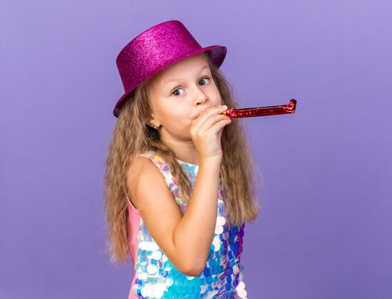 帽子可爱的金发小女孩戴着紫罗兰色的派对帽吹着派对哨子 在紫色的墙壁上看着与世隔绝的复制空间金发孤立哨子