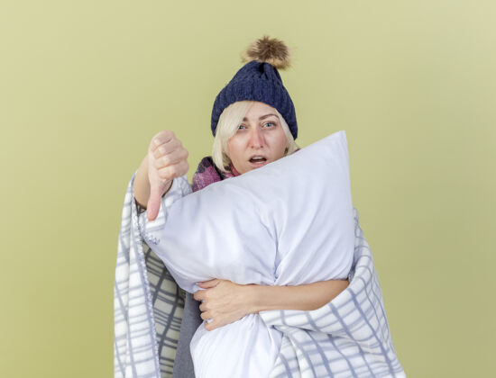 脸年轻的金发生病的女人戴着冬天的帽子 围着格子呢围巾 大拇指朝下 抱着一个孤立的枕头在橄榄绿的墙上持有枕头向下
