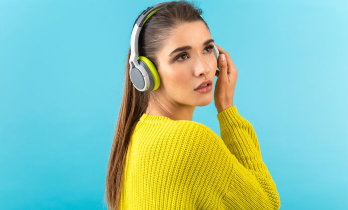 女人戴着无线耳机听音乐的女人开心地穿着黄色针织毛衣摆着蓝色的姿势无线耳机时髦