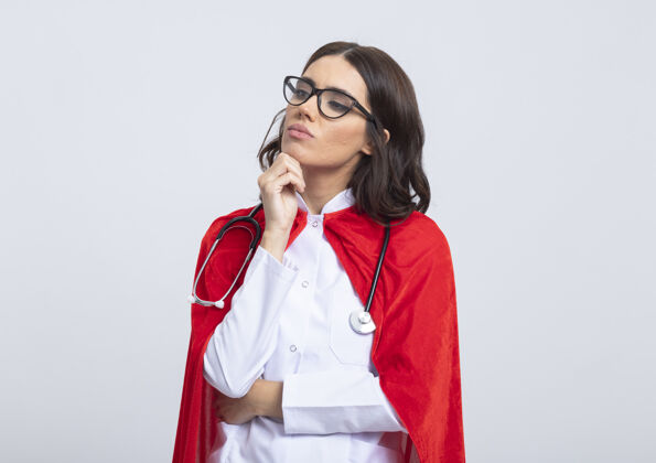 光学自信的女超人穿着医生制服 戴着红色斗篷和听诊器 戴着眼镜 把手放在下巴上 看着隔离在白墙上的一面超级英雄下巴眼镜