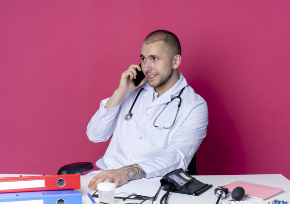 微笑面带微笑的年轻男医生穿着医用长袍 听诊器坐在办公桌旁 手放在办公桌上 手放在粉红色的墙上 手拿着工作工具 一边打电话男性姿势桌子