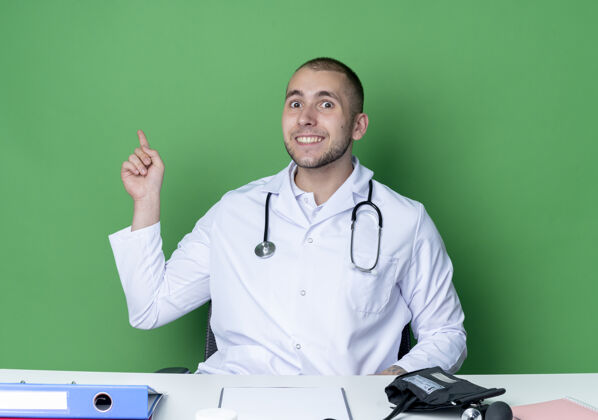 医疗面带微笑的年轻男医生穿着医用长袍和听诊器坐在办公桌旁 工作工具被隔离在绿色的墙上感觉穿着表情