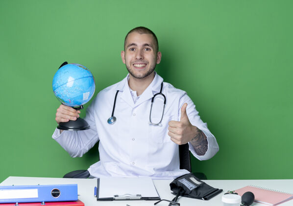 表情面带微笑的年轻男医生穿着医用长袍 听诊器坐在办公桌旁 手持工作工具 举着地球仪 在绿色的墙上孤立地竖起大拇指抱着表演穿着