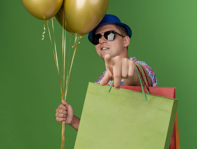 蓝色高兴的年轻人戴着蓝色的帽子和眼镜 拿着气球和礼品袋 向你展示绿色隔离的姿态帽子穿包