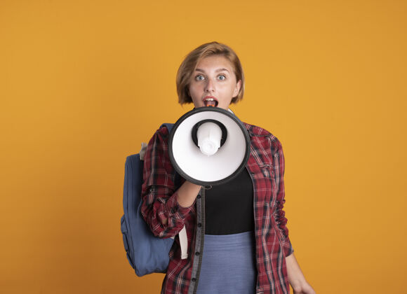 学生惊讶的年轻女学生戴着耳机背着书包对着橙色墙上的扬声器大声说话扬声器年轻人女性