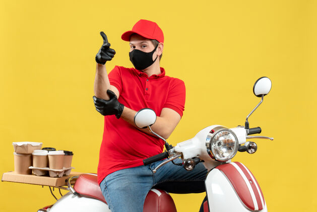摩托车俯视图：穿着红色上衣 戴着帽子 戴着手套 戴着医用面罩 坐在黄色背景的踏板车上传递命令 心满意足 雄心勃勃的年轻人黄色人男人