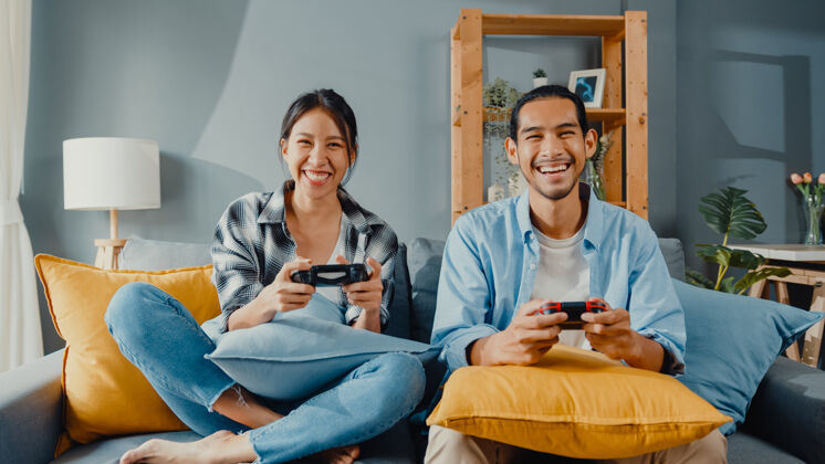 一起快乐的亚洲年轻夫妇男女坐在沙发上玩游戏手柄控制器游戏人房子