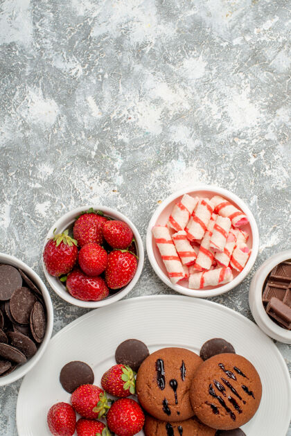 水果顶视饼干草莓和圆形巧克力在白色椭圆形盘子包围着碗糖果草莓和巧克力在灰白色的桌子底部食品巧克力盘子