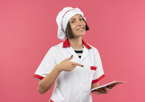 垫身着厨师制服的年轻女厨师面带微笑 手持并指着隔离在粉红色墙上的便笺本衣服脸人