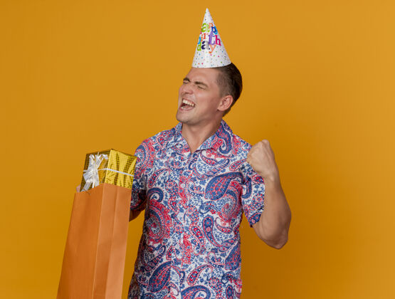 是快乐的年轻人闭着眼睛 戴着生日帽 拿着礼品袋 在橙色上显示“是”的手势手势生日包