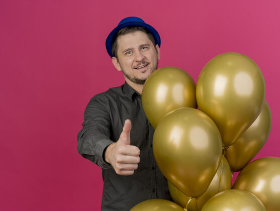 帽子高兴的年轻人戴着蓝色的帽子站在气球旁边 拇指向上 孤立地站在粉红色的气球上拜托小伙子派对
