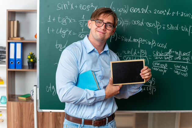 自信年轻的男老师戴着眼镜拿着书拿着小黑板看着相机微笑着自信地站在教室里数学公式的黑板边数学教室黑板