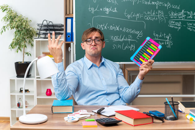 账单年轻的男老师戴着眼镜 带着账单 在教室的黑板前拿着书和笔记坐在课桌旁备课教室准备坐着